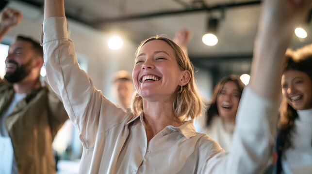 A woman beams with joy, her arms raised in a gesture of triumph, surrounded by a blur of equally jubilant people celebrating in an indoor setting.
