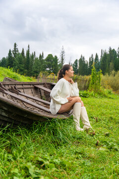 Young woman in white sweater and knee-high boots sits on an old wooden boat, surrounded by lush green grass and trees in a natural outdoor setting