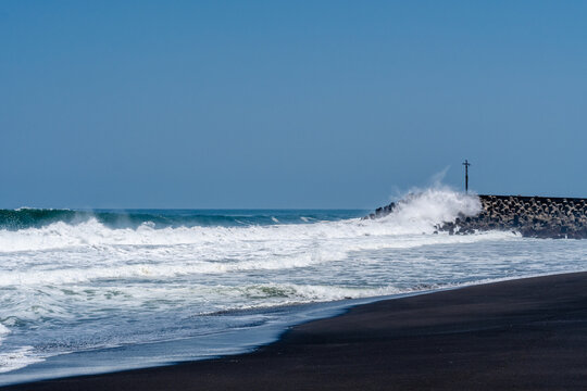Large ocean waves crashing against a concrete breakwater structure on a black sand beach