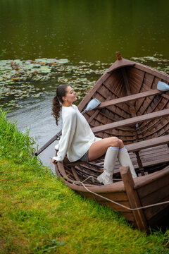 Young woman in white sweater and knee-high socks sitting on wooden boat by tranquil lake surrounded by lush greenery and lily pads
