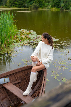 Young woman relaxing in wooden rowboat by lake on autumn day. Cozy sweater and rubber boots represent seasonal outdoor lifestyle and slow living in nature