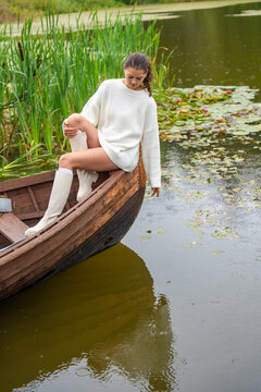 Young woman in white sweater sits on wooden boat, reaching towards water surface surrounded by lily pads and lush greenery in serene lake setting
