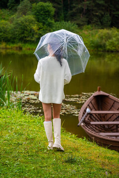 Young woman in white sweater and knee-high socks walks with a clear umbrella by a tranquil lake, wooden boat moored nearby, surrounded by lush greenery
