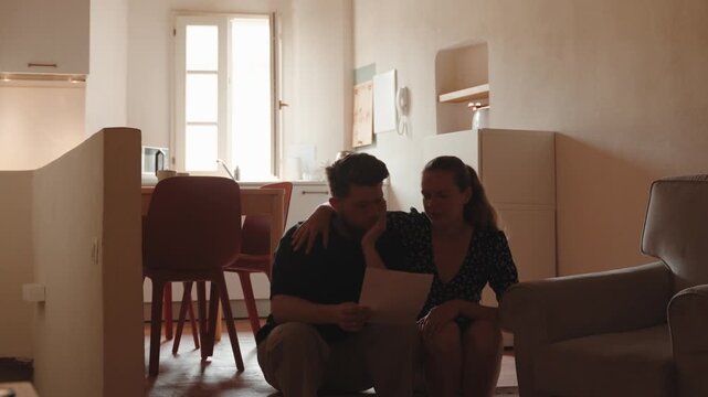 Worried couple receiving bad news in a letter, experiencing financial difficulties, debt problems, and stress while reading a document together in their apartment