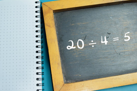 Chalkboard with phrase 20 &divide; 4 = 5 math division equation on wooden desk. For basic arithmetic and school education