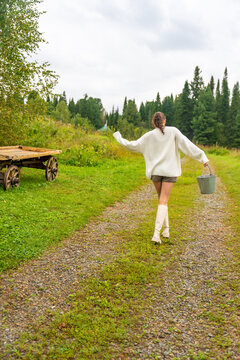 Young woman in white sweater and shorts walks along a gravel path carrying a bucket, with a wooden cart and trees in the background
