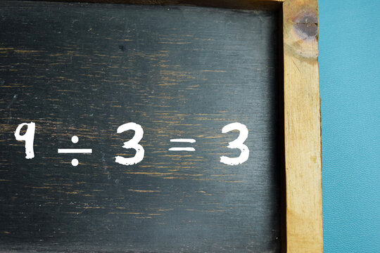 Chalkboard with phrase 9 &divide; 3 = 3 division equation on wooden desk. For school education and learning