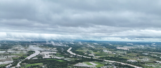 Fototapeta premium Rain clouds blanket swollen river winding through lush agricultural fields, dense mist rolls across the flooded rural landscape in gloomy monsoon day.