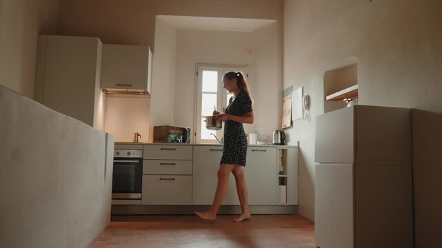 Barefoot young woman in a dress cooking and preparing food in a cozy, bright kitchen, enjoying her daily routine and healthy lifestyle at home in her apartment