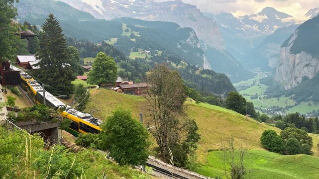 Yellow mountain train crosses the green hillside near Wengen Switzerland, Rail travel footage overlooks chalets and the deep Lauterbrunnen Valley