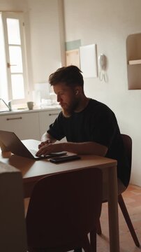 Focused bearded freelancer working remotely on a laptop with wireless earbuds, typing at a kitchen table while listening to music or a podcast in his cozy home office