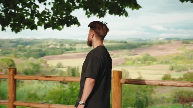 Young bearded man walking along wooden fence admiring the beautiful green rolling hills and valleys of tuscany, enjoying a peaceful day on his italian holiday