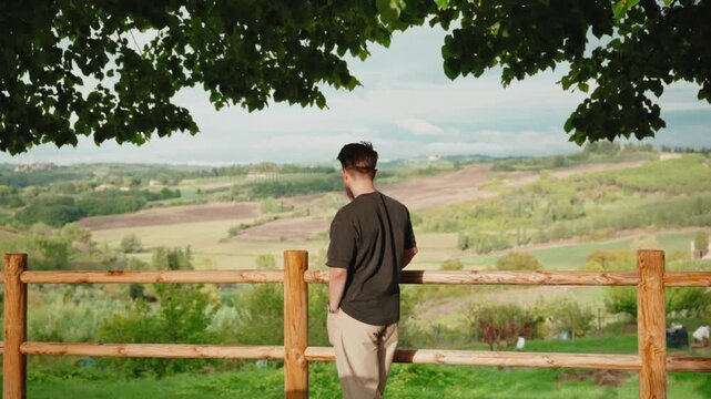 Young man leaning on a wooden fence enjoying the beautiful panoramic view of the tuscan countryside and contemplating the idyllic green landscape from a hill