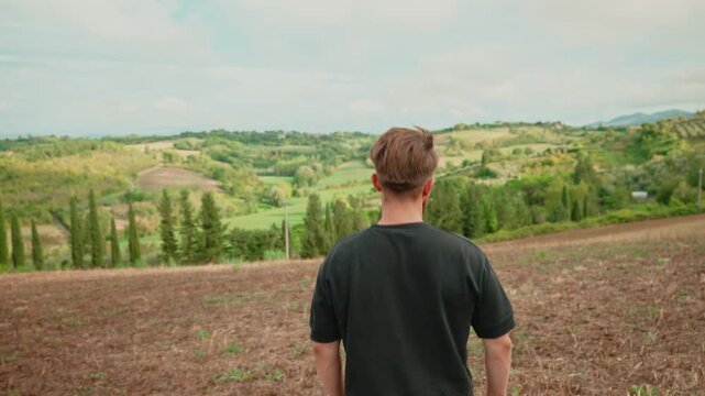 Young man standing in a field looking at the stunning tuscan landscape with cypress trees, rolling hills, and green vineyards in the idyllic italian countryside