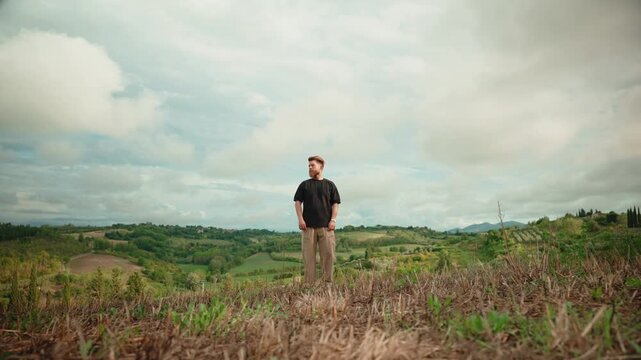 Young man standing alone in a vast tuscan field, contemplating the rolling green hills and cloudy sky, embodying freedom, solitude, and connection with nature in italy