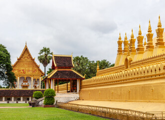 Fototapeta premium Golden stupa and temple complex stand under overcast sky, surrounded by green lawn and paved walkway in Vientiane, Laos.
