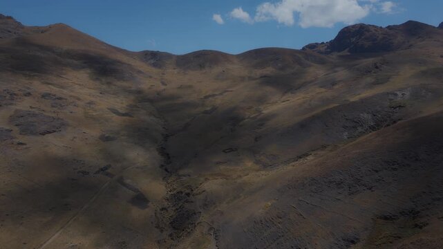 Aerial view of dry, rolling mountain slopes and ravines in the Peruvian Andes highlands, with cloud shadows sweeping across the vast arid mountain landscape.