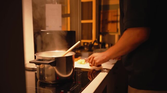 Young man preparing dinner in a warm and cozy kitchen at home, chopping vegetables on a cutting board while water boils in a steaming pot on a modern induction stove