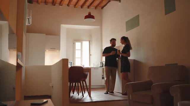 Romantic young couple enjoying morning coffee, talking and smiling together in their cozy sunlit apartment, sharing an intimate moment of connection and affection