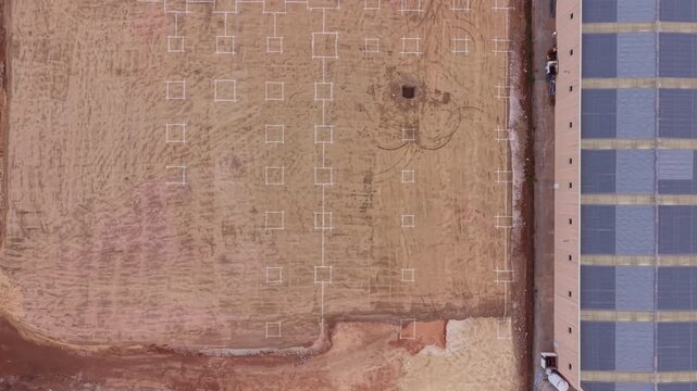 Top-down view of a datacenter construction site showing marked foundation grid on bare earth beside an existing building with solar panels on its roof