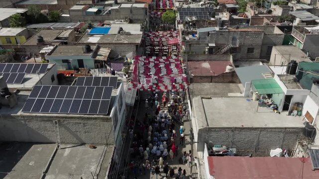 Drone flight above flat rooftops and solar panels in San Juan de Ocotan, Jalisco, looking down a narrow street strung with pink paper banners where riders move through the crowd. Aerial flyover shot