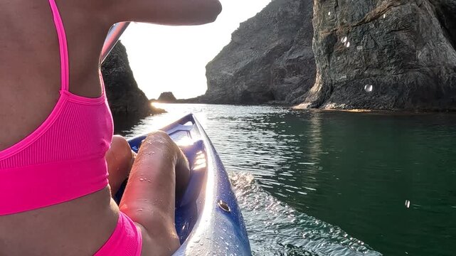 Pink bikini woman paddling blue kayak towards rocky cliffs during a bright golden sunset. Over-the-shoulder point of view shot.