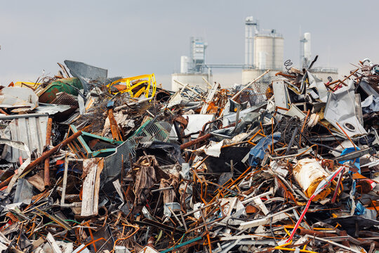 Massive concrete silos tower behind piles of scattered scrap metal and industrial debris. Scene shows impact of heavy manufacturing on fragile environment, urging sustainable waste solutions