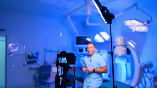 Camera records a video in the medical room with blue light. Male medic leaning on the operating table talks to camera. Blog content creation.
