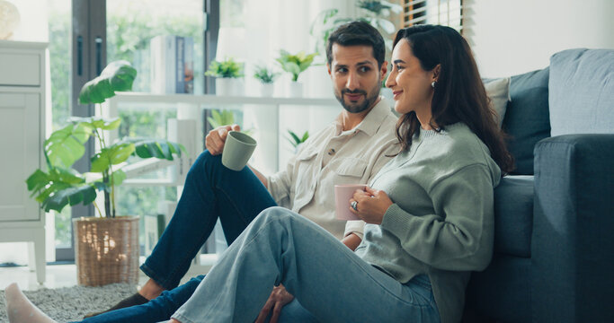 Happy young adult Hispanic couple drinking coffee and relaxing sit on floor in living room at home. Domestic lifestyle, positive weekend leisure, cheerful relationship bonding, family love concept.