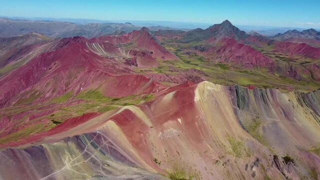 Aerial view of the colorful mineral-rich slopes of Vinicunca, featuring vibrant red, yellow, and turquoise sediment layers under a clear blue sky in Quispicanchi Province, Cusco, Peru.