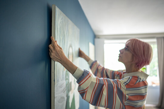 Concentrated woman painter decorating apartment, hanging own canvas artwork on wall, focused female carefully putting and aligning large picture on wall in room, interior improvement and creativity.