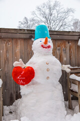 Cute funny snowman holding red heart toy outdoors near rustic wooden wall, winter love, Valentine's day romance and holiday seasonal greetings concept © Katharina