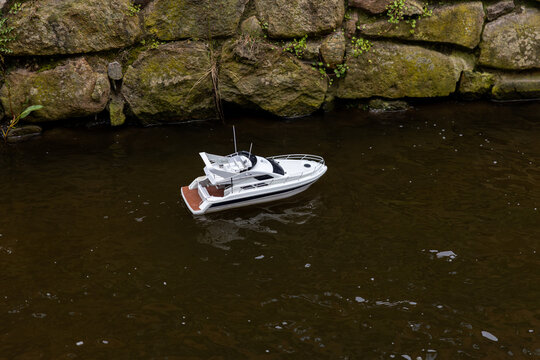 Remote control model boat moves along narrow canal creating ripples stone embankment. Outdoor hobby scene highlights miniature technology calm water surface, urban textures focused recreational play