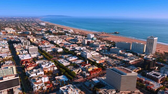 Santa Monica cityscape and coastline from above. Streets and buildings extend toward a pier and sandy beach beside the blue Pacific Ocean.