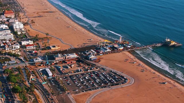 Santa Monica Pier with amusement rides and beach parking. A Ferris wheel and roller coaster stand above the water beside a broad beach and rows of parked cars.
