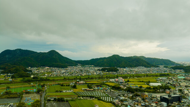 Aerial view of Mishima City featuring golden rice fields, residential neighborhoods, and lush green mountain ranges under a cloudy sky in Mishima, Shizuoka, Japan.