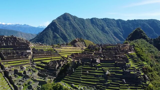 Aerial view of the ancient Inca citadel of Machu Picchu with stone terraces, historic ruins, and lush green mountains under a clear blue sky in Urubamba Province, Cusco, Peru.