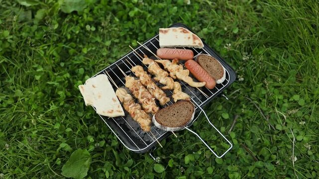 Variety of food including chicken skewers, sausages, burger patties, and flatbread grilling on a wire rack of a small black charcoal grill. Preparing a delicious mixed grill lunch during an outdoor
