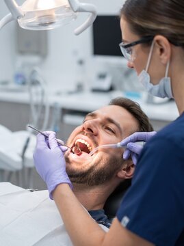 A bearded man lies back with his mouth open while a dentist examines his teeth with a mirror and suction tube during a dental procedure.