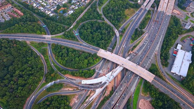 Highway interchange with looping ramps and intersecting bridges. Roads curve through wooded areas around a divided highway and an unfinished bridge section.