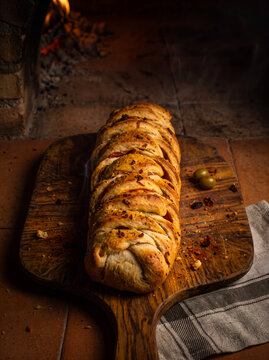 Freshly baked braided stuffed bread on a rustic wooden board in front of a wood-fired stone oven; moody lighting, vertical view