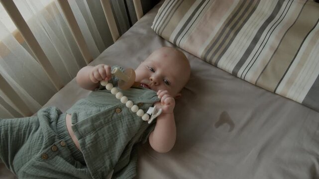 Portrait of Caucasian infant lying in crib, holding pacifier teether and looking at camera. Footage suitable for parenting campaigns, pediatric healthcare content and baby product advertising