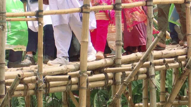 A population of people of different ages is crossing a temporary bamboo footbridge over the river