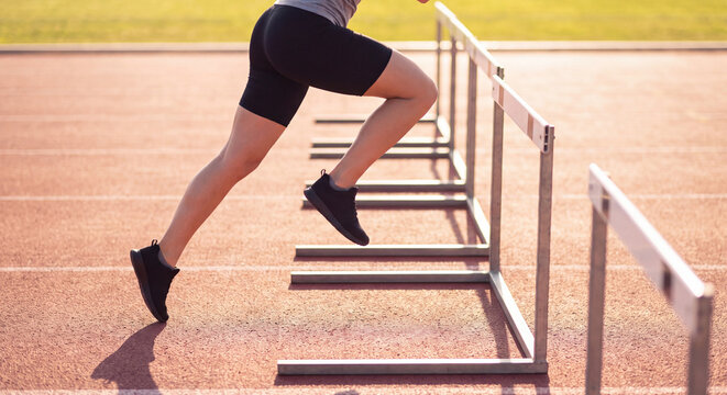 Low angle close up of legs of an athletic runner leaping over hurdle obstacles on red running track outdoors.