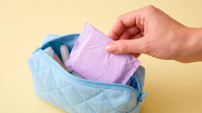 Hand placing a purple sanitary pad into a light blue quilted cosmetic bag on yellow background