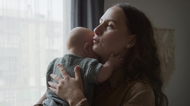 Caucasian mother comforting and holding fussy infant baby near window. Visual asset fitting parenting education, maternal healthcare campaigns, and baby care commercials