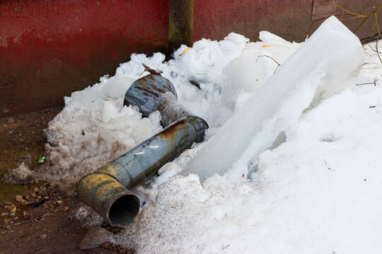 Fallen metal drainpipe lying in snowbanks with massive ice icicles near a residential wall during the cold early spring season, danger of ice falling from roofs