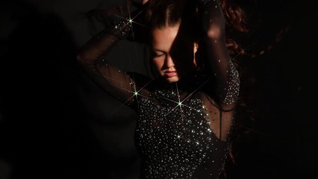 Fashion editorial portrait, woman in a rhinestone gown at a dark wall. Direct gaze, hard spotlight and deep shadow for a lookbook or casting card.