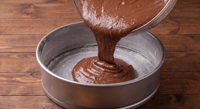 Thick dark chocolate cake batter being poured from a glass bowl into a metal springform baking tin