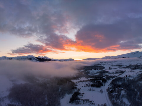 Aerial view of the snowy winter landscape on Kvaloya island, featuring rolling hills covered in snow, low-hanging clouds, and a vibrant sunset sky in Troms, Norway.
